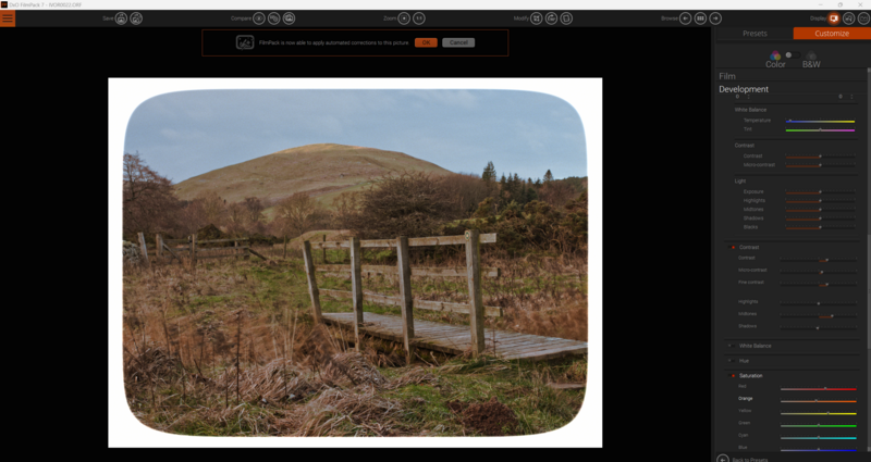 Wooden fence gate in overgrown field with rolling hill and evergreen trees in background.