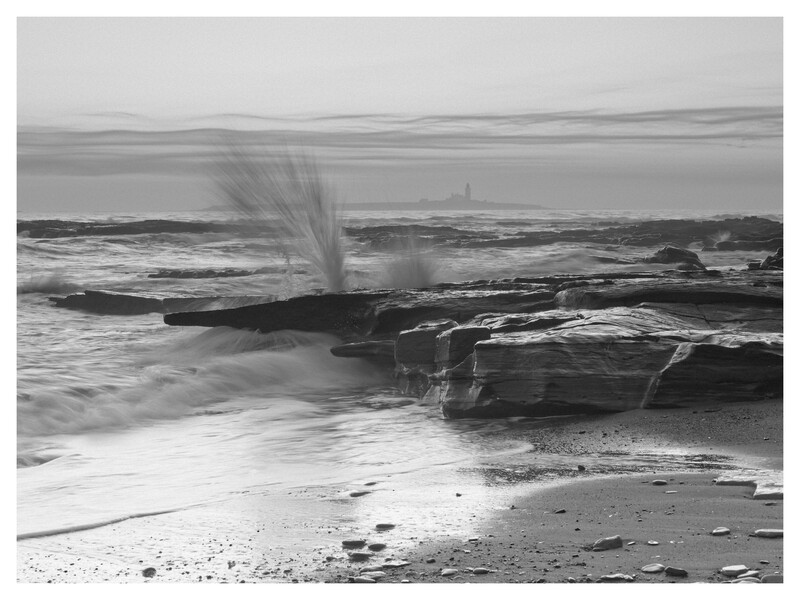 Wave crashes against rocky coastal outcropping, spraying water high into the air on an overcast day.