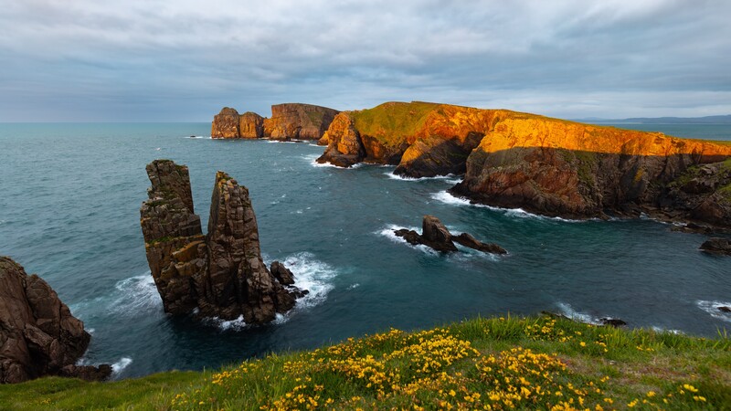 Dramatic coastal cliffs with golden-lit rock formations and columnar basalt stacks rising from turquoise ocean waters.