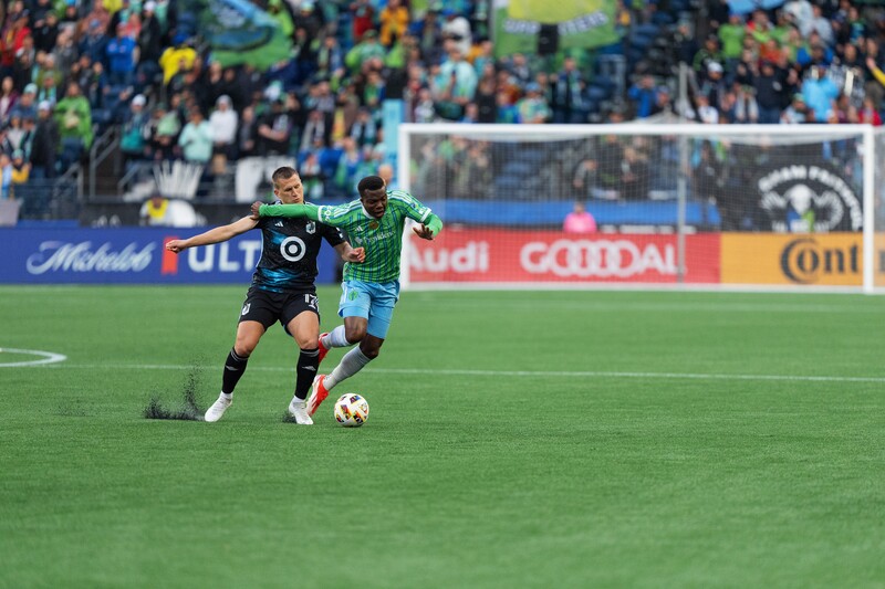 Soccer player in green jersey controlling the ball while being defended by player in dark jersey during professional match.