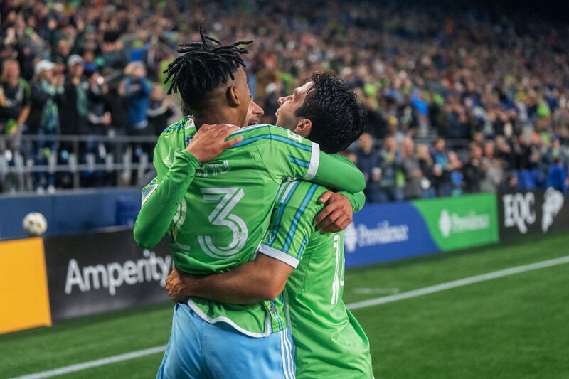 Two soccer players in bright green uniforms embrace while celebrating on the field in front of a large crowd.
