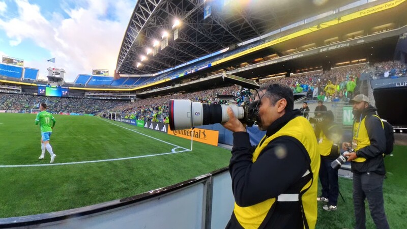 Photographer with telephoto lens at stadium sideline during soccer match.