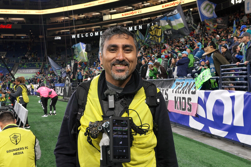 Sports photographer wearing yellow and black gear holds professional video camera at indoor stadium event.