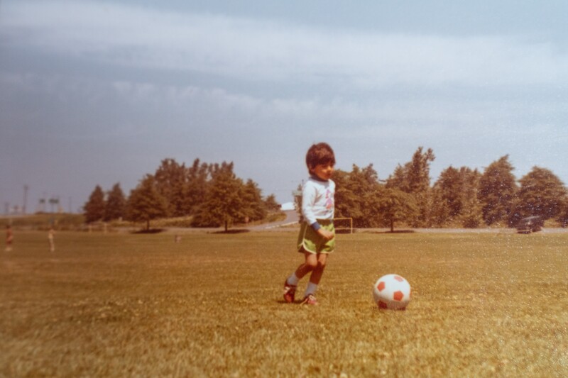 Young child kicking a soccer ball across an open field with coniferous trees in the background.