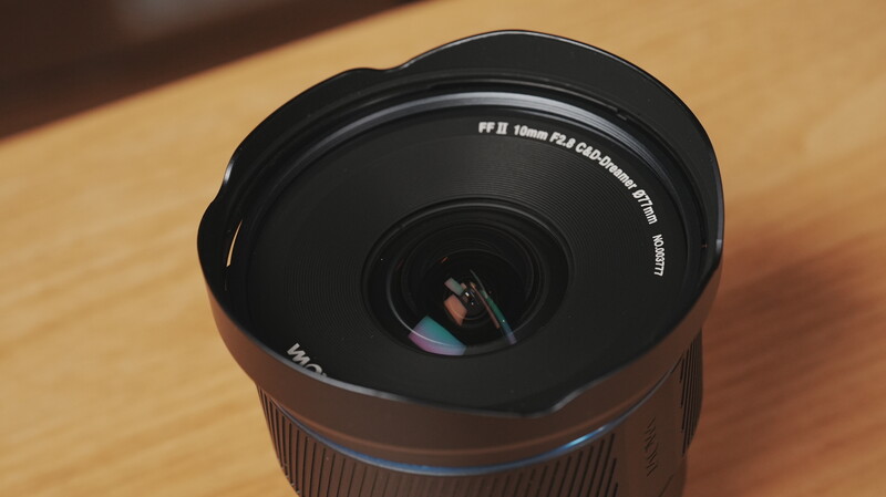 Wide-angle lens with front element cap removed, showing internal glass elements and aperture mechanism on wooden surface.