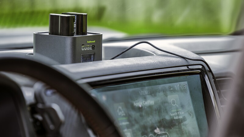 Dashboard-mounted dashcam with external battery pack positioned on car windshield against blurred green background.