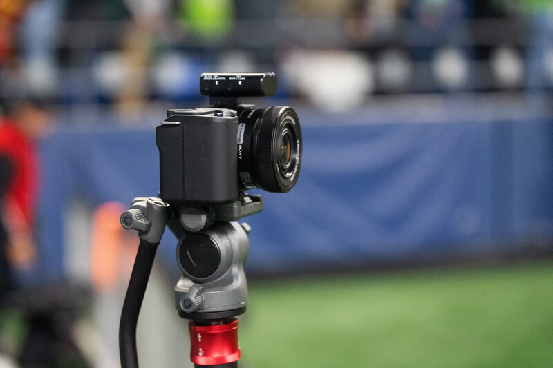 Camera mounted on a fluid head tripod, positioned to film a sports event with stadium and crowd in soft focus background.