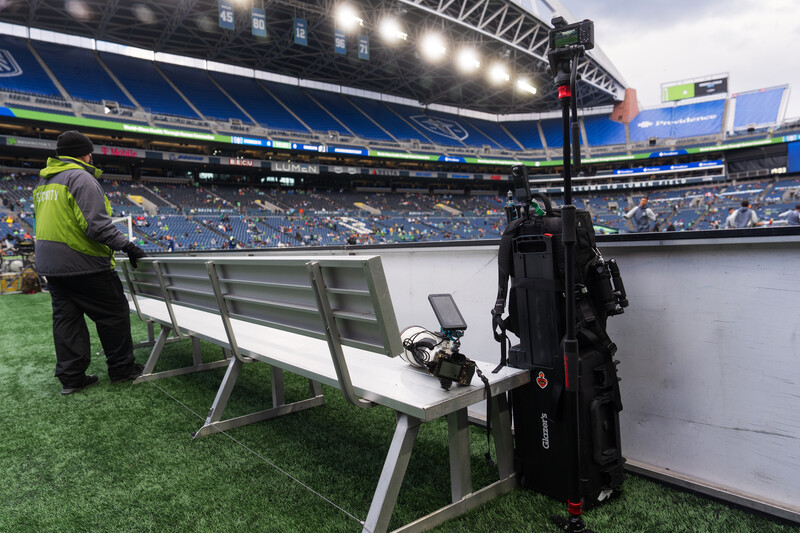 Camera operator with telephoto lens on stadium sideline during daytime match.