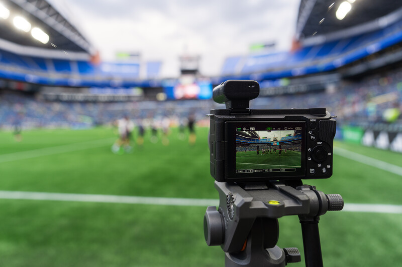 Camera on tripod recording video of soccer match in large stadium with blue seating.