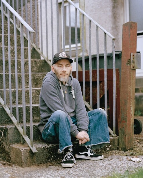 Young person in cap and hoodie sitting against a wooden fence gate, looking at camera.