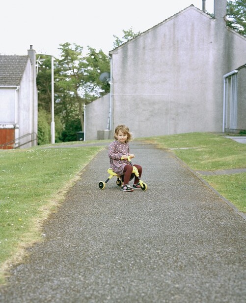 Young child riding a tricycle down a narrow asphalt path between concrete structures and grass.
