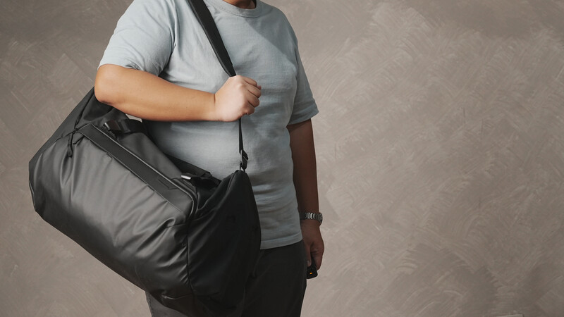 Person wearing a gray t-shirt carrying a large black duffel bag across their torso.