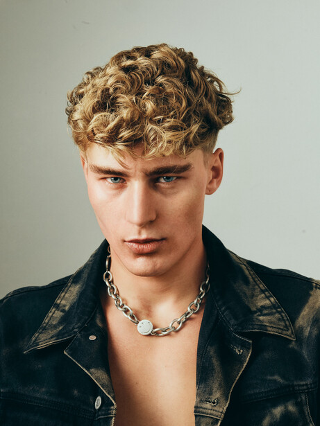 Portrait of a young man with blonde curly hair wearing a black leather jacket and chain necklace against a neutral background.