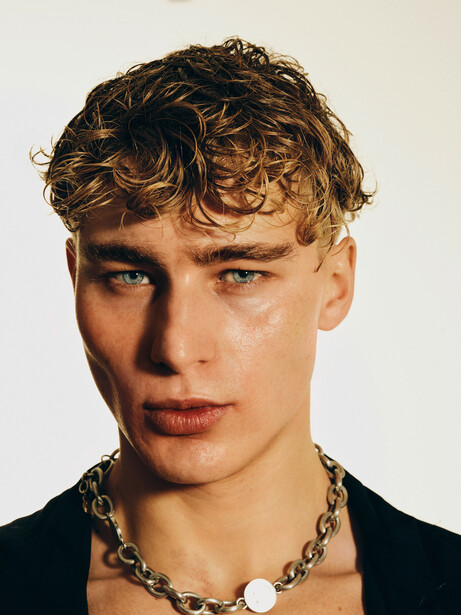 Young man with curly blonde hair wearing black shirt and chain necklace against neutral background.