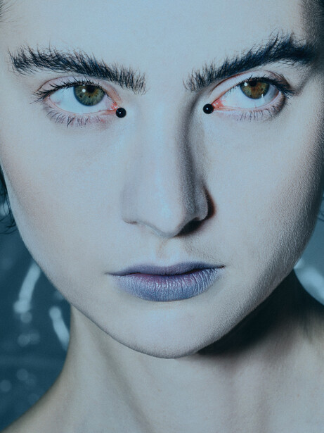 Close-up portrait of a person's face with pale complexion, dark eyebrows, and blue eyes with subtle makeup details.