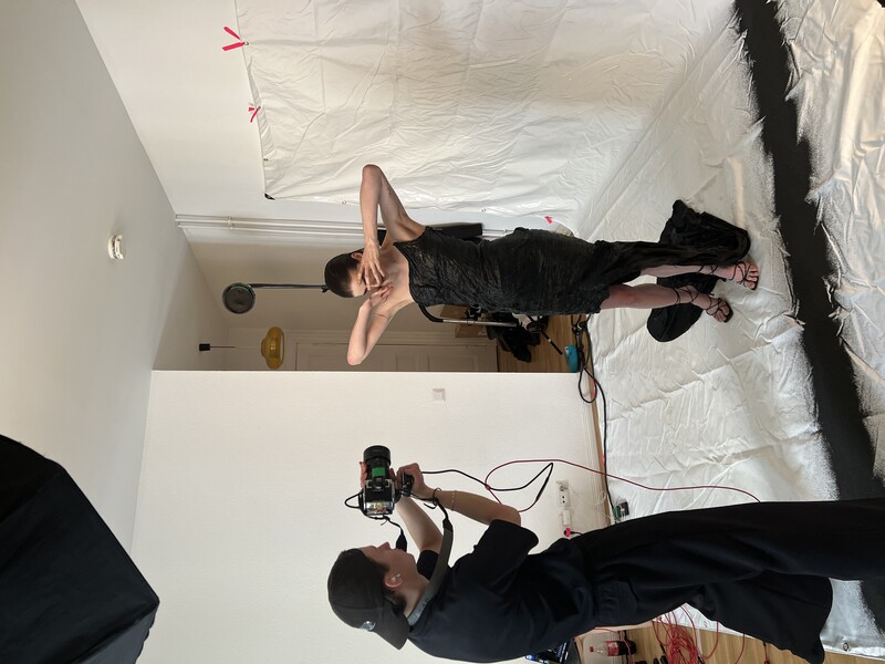 Photographer shooting a portrait of a shirtless male model posing against white backdrop with studio lighting setup.
