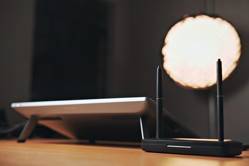 Black Wi-Fi router with two antennas positioned next to a tablet on a wooden desk with warm bokeh lighting in the background.