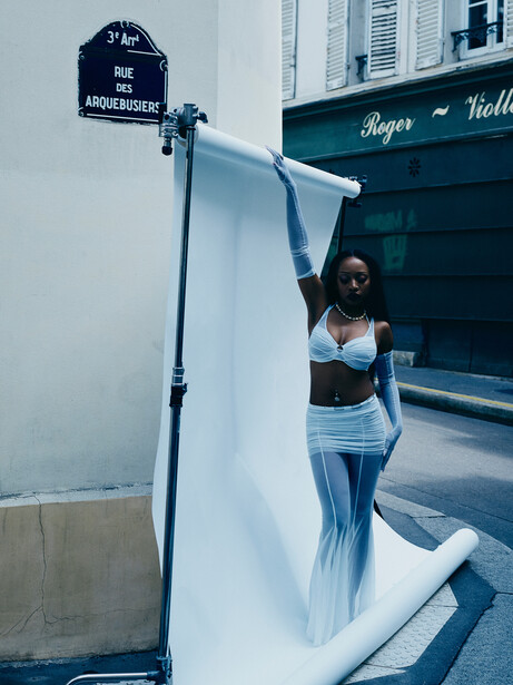 Woman posing against white fabric backdrop on urban street with storefront behind her.