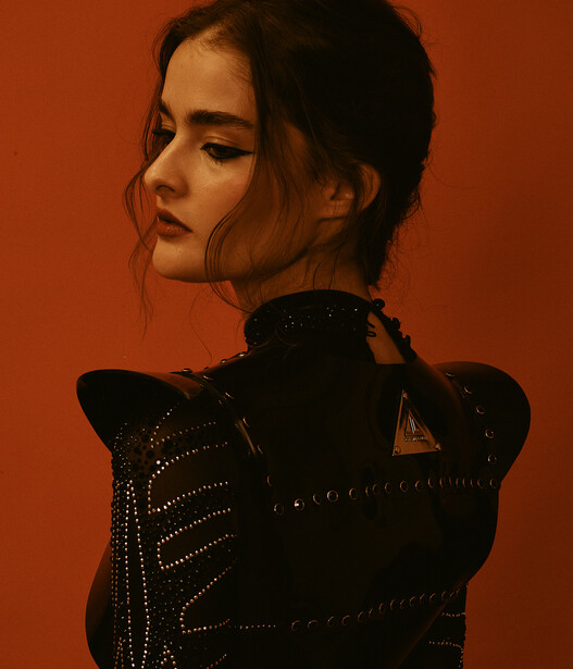 Woman in studded black jacket photographed in profile against warm orange background.