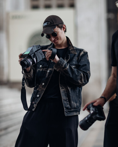 Photographer in denim jacket and cap holding DSLR camera with another person partially visible on right.