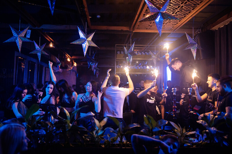 Energetic crowd at indoor event with blue and white star decorations overhead and warm stage lighting.