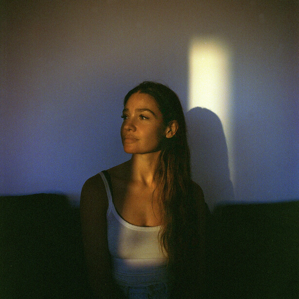 Woman with long dark hair sitting against a blue wall, lit by warm window light creating dramatic shadows.