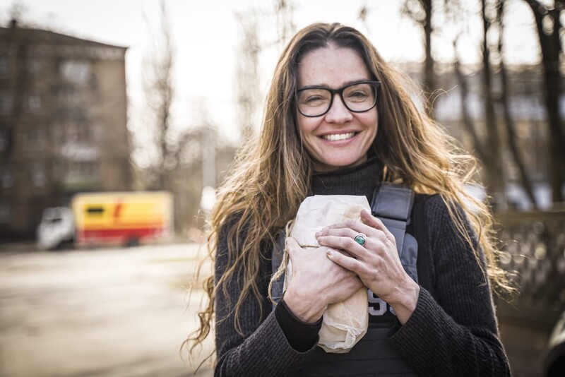 Woman wearing glasses and dark sweater smiling at camera while holding a small object outdoors.