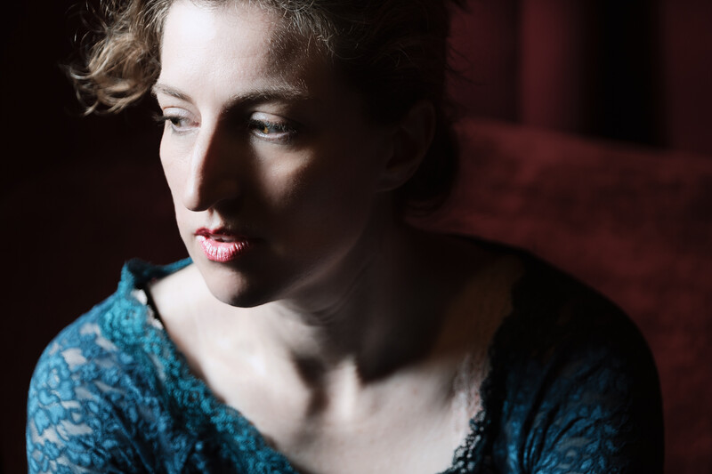 Woman with curled hair wearing a teal patterned top, photographed against a dark red background with dramatic side lighting.