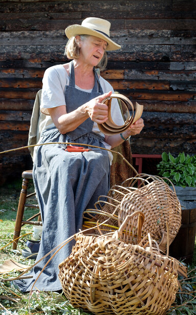 Craftsperson seated against a wooden wall, holding woven baskets and demonstrating traditional basketry work.