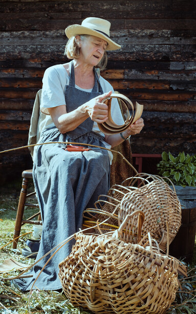 Craftsperson in overalls and straw hat holding woven baskets against a rustic wooden backdrop.