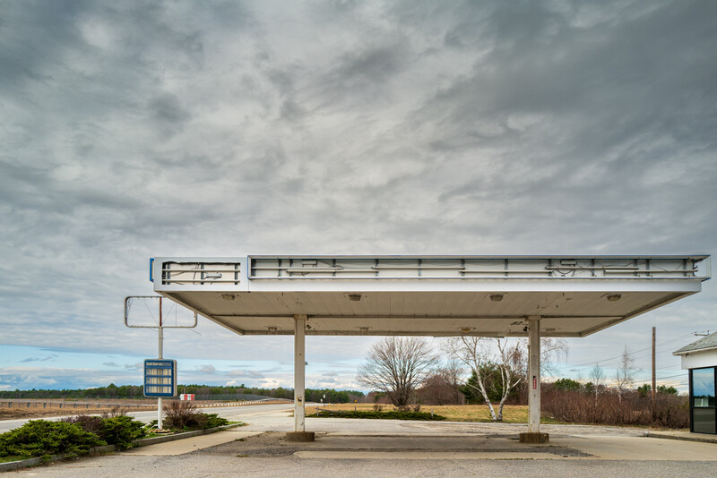Abandoned gas station with a large canopy structure under overcast sky and bare trees.