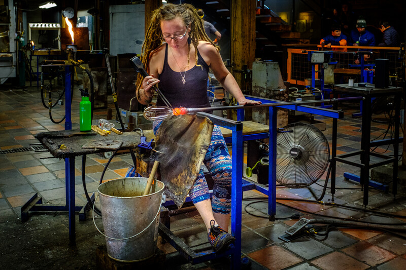 A person with dreadlocks wearing glasses works on metal fabrication in a workshop, holding a glowing piece in metal-working tongs over a bucket.