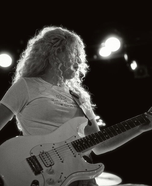 Musician with long curly hair playing electric guitar on stage with stage lights in background.
