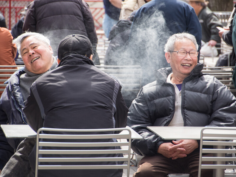 Two elderly people sitting on bleachers in cold weather, their breath visible as vapor in the air.