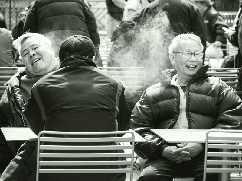 Two smiling women seated outdoors on a bench in winter clothing, with their breath visible in cold air.