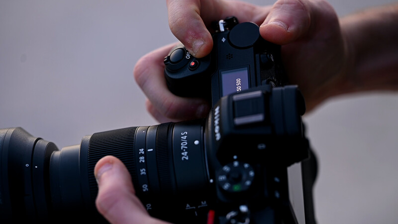 Hands holding a mirrorless camera with attached lens, viewed from above against a neutral gray background.