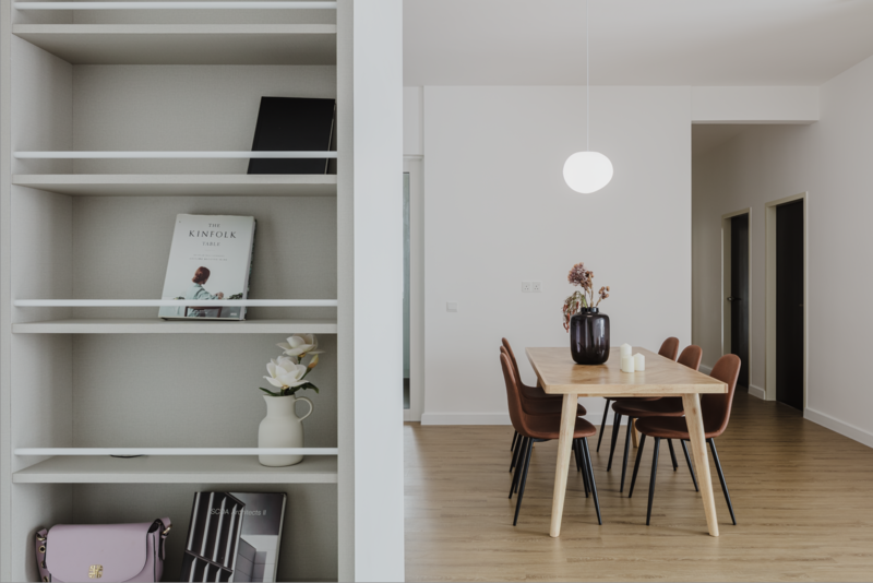 Modern minimalist interior with open shelving on left displaying decorative objects and a dining area with wooden table and chairs on right.