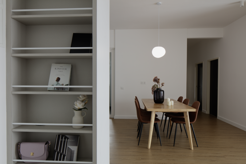 Modern minimalist interior with open shelving unit displaying decor and a dining area with wooden table and chairs.