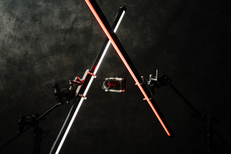 Two neon tubes crossed in an X formation against a dark textured background, with studio lighting equipment visible.