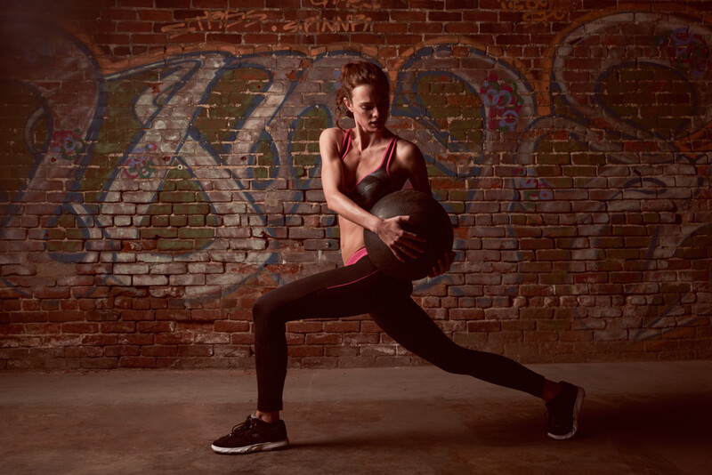 Athlete performing a deep lunge stretch against a brick wall with large graffiti art.