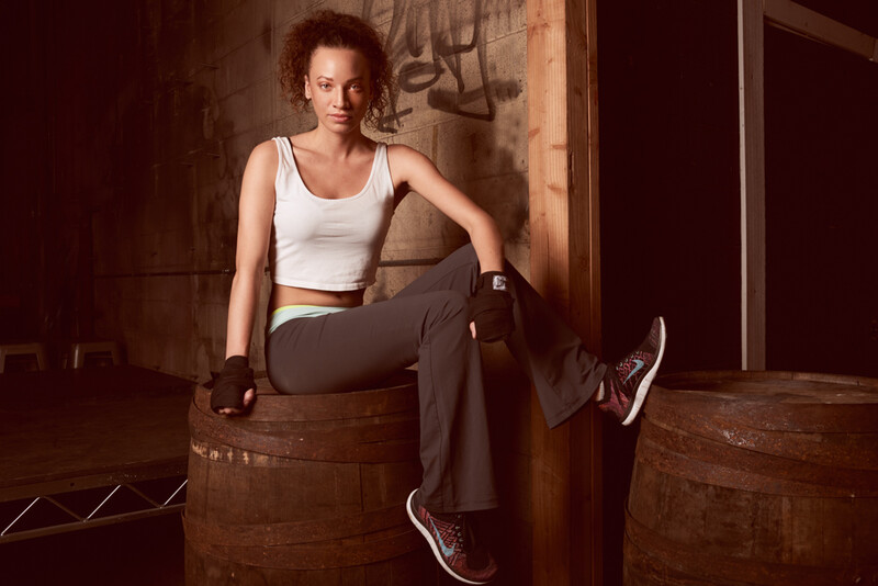 Woman in white crop top and dark pants sitting on wooden barrel, wearing boxing gloves.