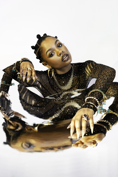 Person with gold-painted skin wearing a black studded jacket and ornate jewelry against a white background.