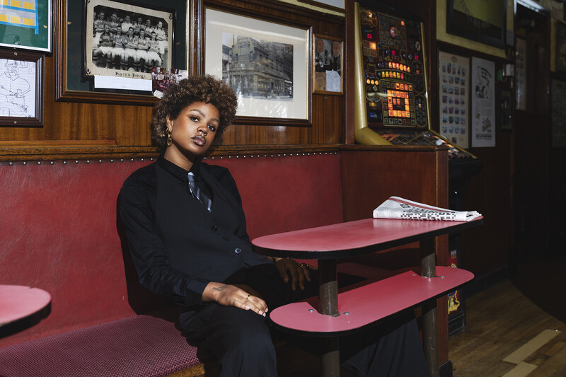 Person in black blazer seated in red diner booth, looking toward camera.