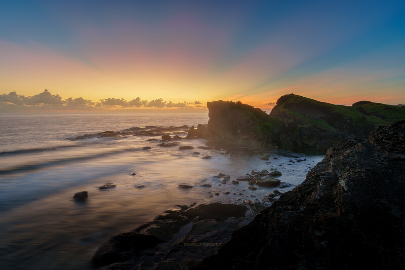 Rocky coastline at sunrise with smooth water and golden sky reflecting on the beach.