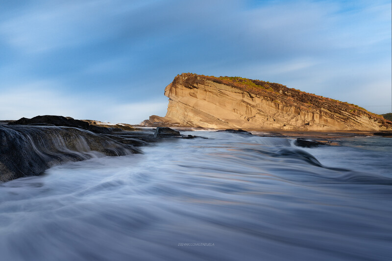 Coastal rock formation with long exposure ocean waves and golden cliffs under blue sky.