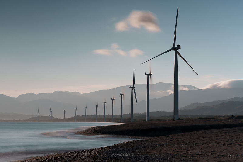 Row of wind turbines on a coastal beach with mountains in the distance under a partially cloudy sky.
