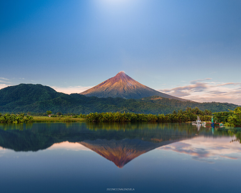 Conical volcano reflected in still lake water at dawn, surrounded by forested slopes.