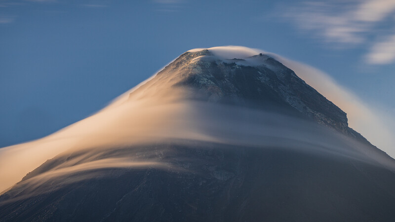Snow-capped volcanic cone rising against blue sky with lenticular cloud formation wrapping around its peak.