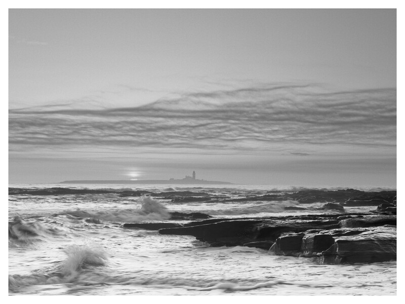 Ocean waves rolling onto a rocky beach at sunset, rendered in black and white with distant lighthouse visible on horizon.