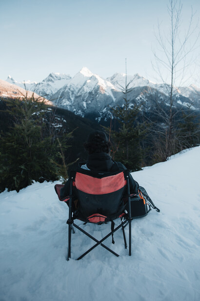 Red and black camping chair on snow with snow-covered mountains in background.